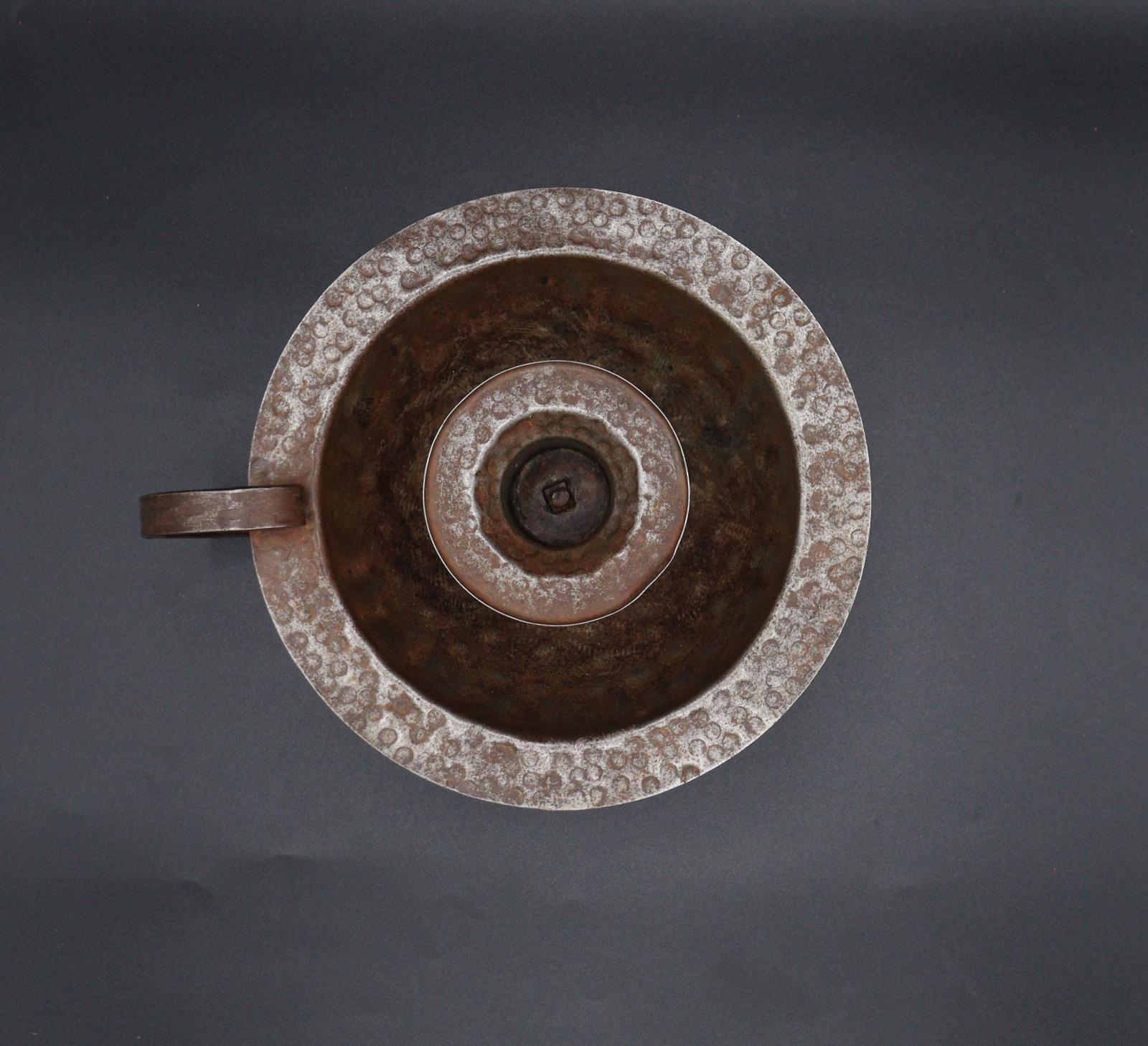 A round, copper-coloured, candle holder, made of tin. It has a bowl for the wax catchment area and a curved handle attached from the rim of the bowl to the base.