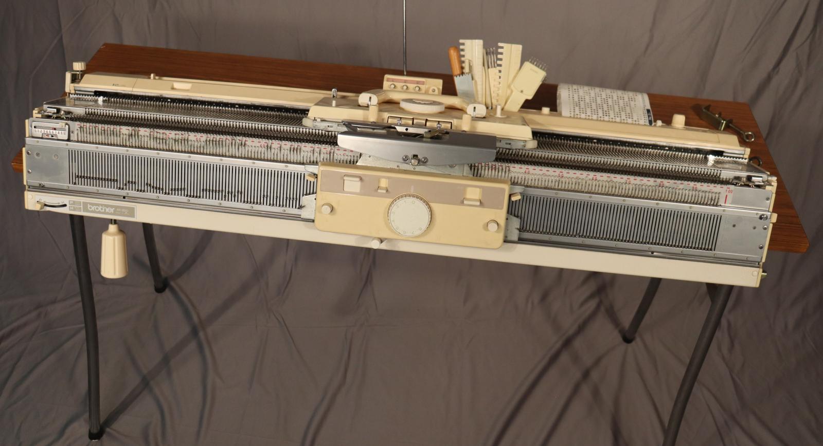 A BROTHER KH-892 Knitting Machine. Made of metal and plastic, approx. a metre in length, situated on a fold out wooden table. Comes with numerous accessories.