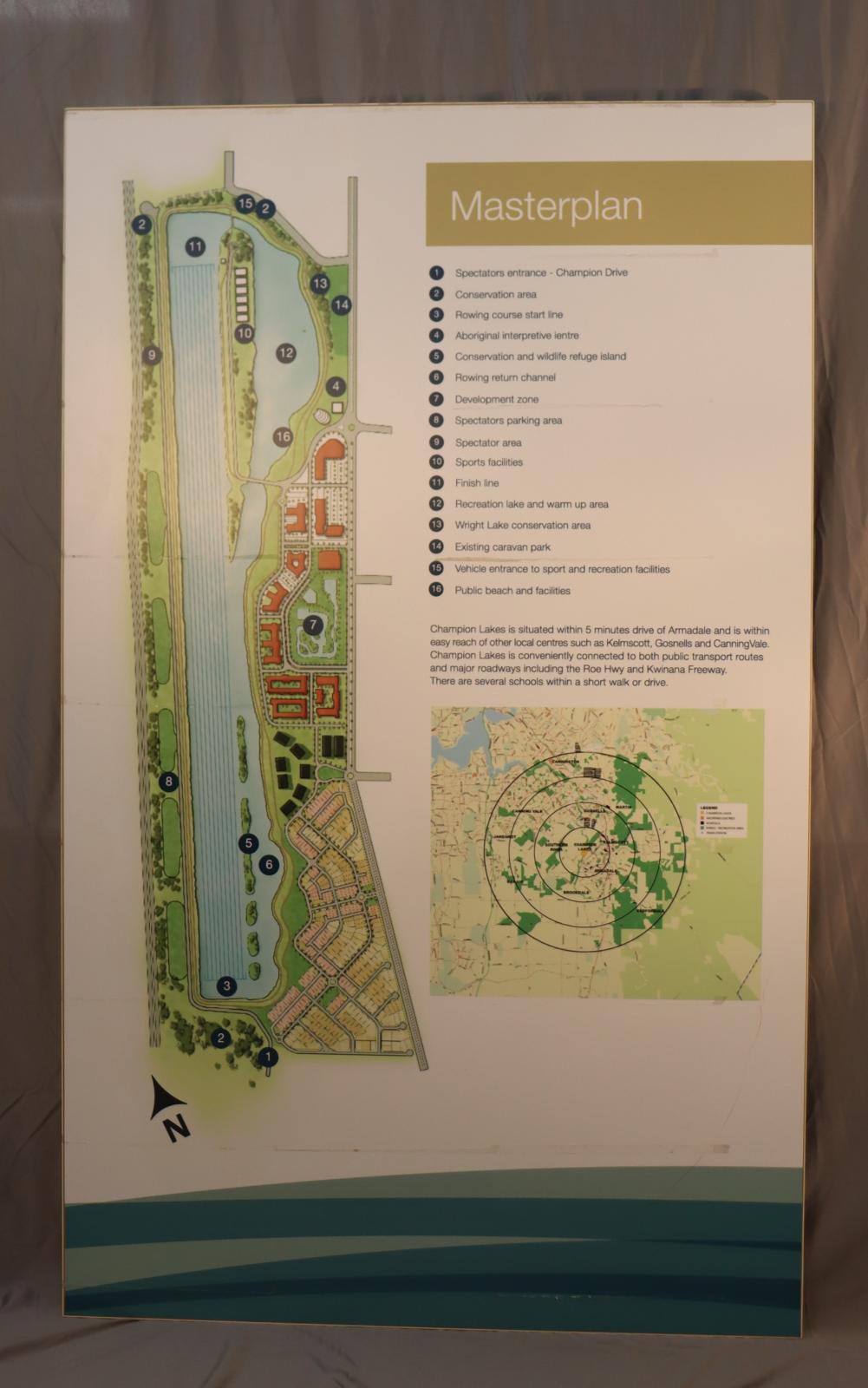 A rectangular sheet of plastic attached to wood. Plastic around the edges of the wood. It is mostly white and light green, green and dark green across bottom. The left half of the picture shows the surrounding areas including residential areas, facilities and lakes and is numbered one to sixteen and marked with a north directional marker.