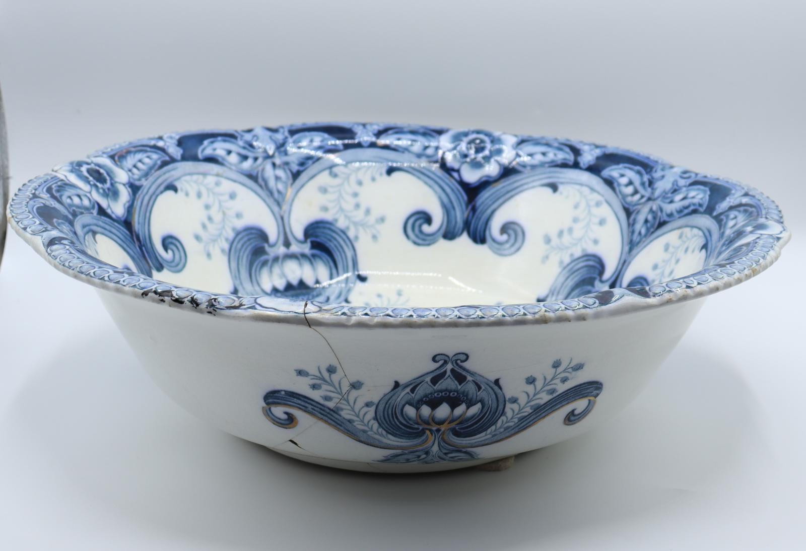 A large, ceramic bowl with a scalloped rim. The inside of the bowl is white, painted with ornate blue detailing. The outside of the bowl is similarly painted with flowers and leaves in three places. Cracks from outside edge down to bottom. There is some black marks on one of the edges.