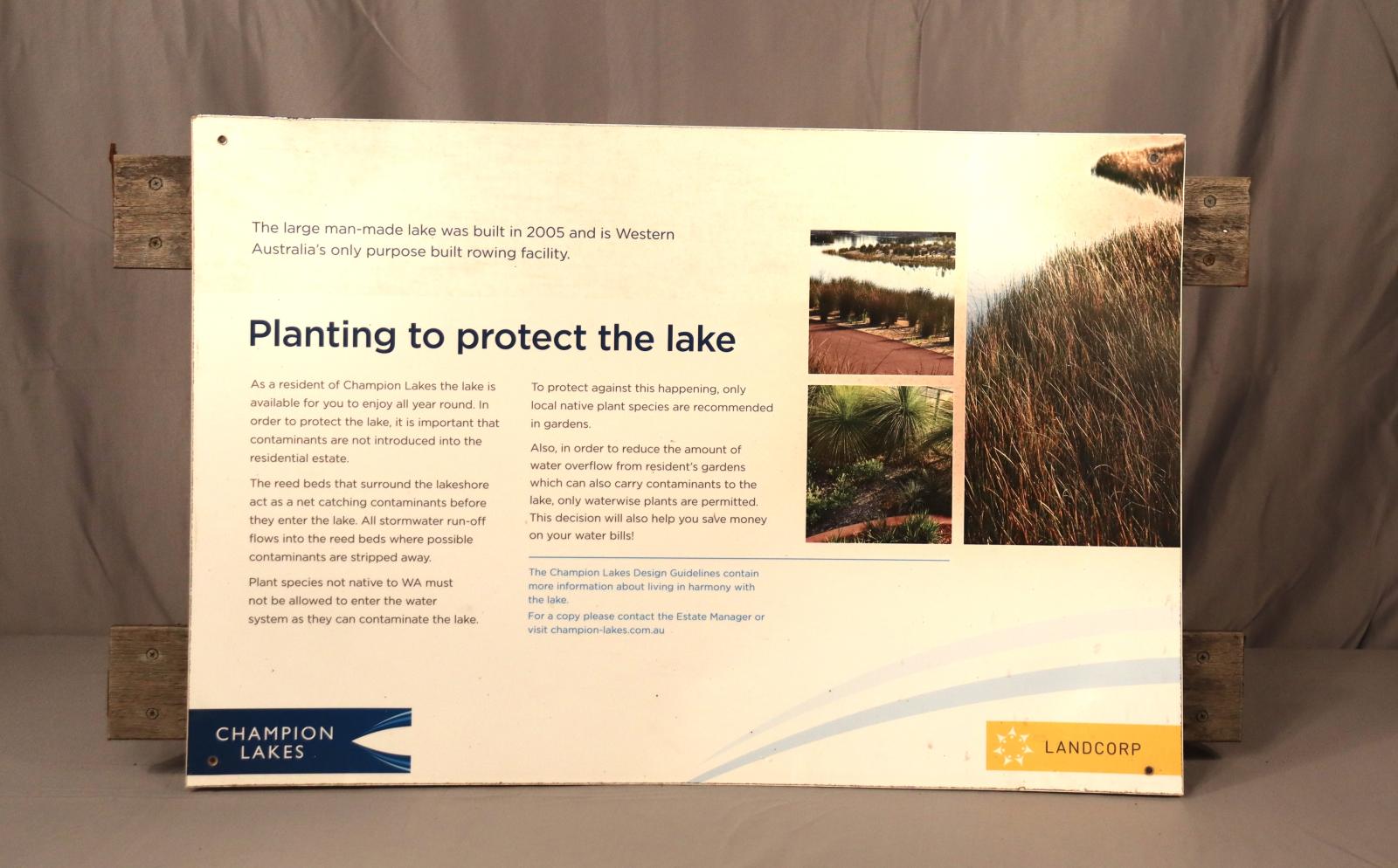 A small cream/white, metal sign, displaying navy-blue text and 3 photos referencing "Planting to protect the lake".