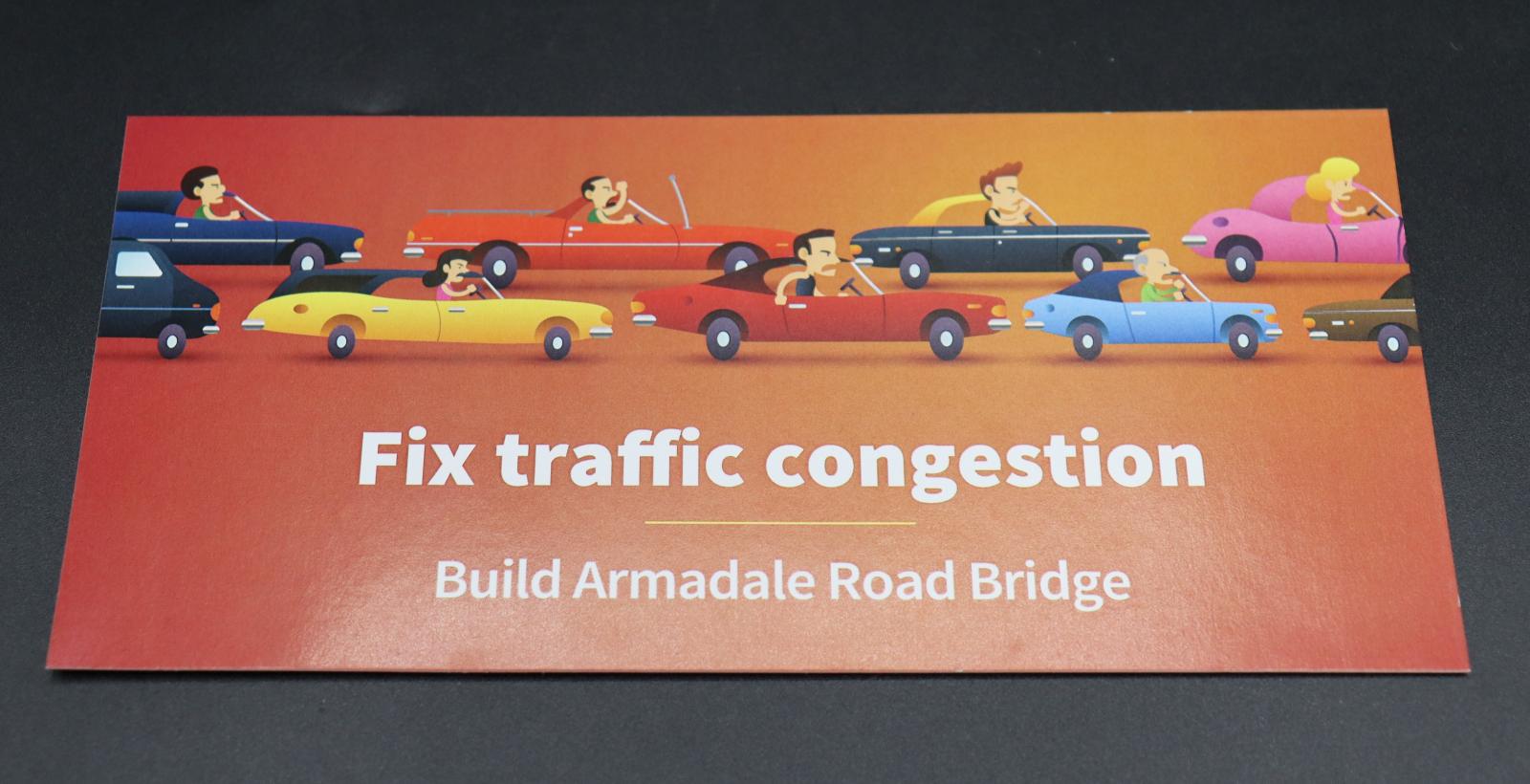 A rectangular, orange, DL (1/3 A4 Size) size flyer. The front reads, in white horizontal text across bottom half, [FIX TRAFFIC CONGESTION / BUILD ARMADALE ROAD BRIDGE]. Above this is an illustration of two lines of cars, facing towards the right with a variety of angry drivers in them.