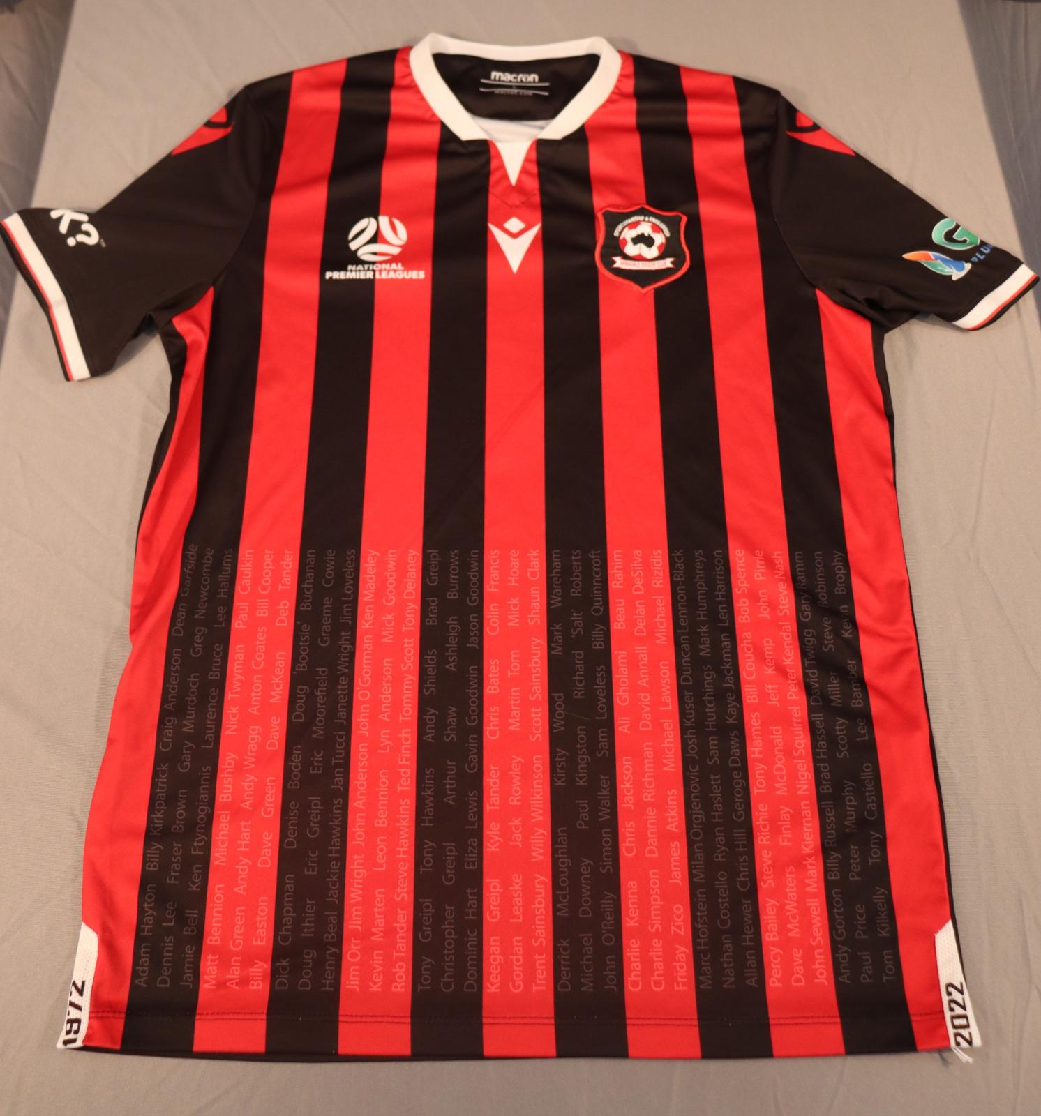 Short sleeve shirt, black and red vertical lines on the front, black sleeves & black back with a white V neckline and white sleeve cuffs. A shield-shaped, black patch with a red edge in top right side of the front of the shirt (left side if worn). On the patch is a red and white round ball with a black image of Australia in the middle.