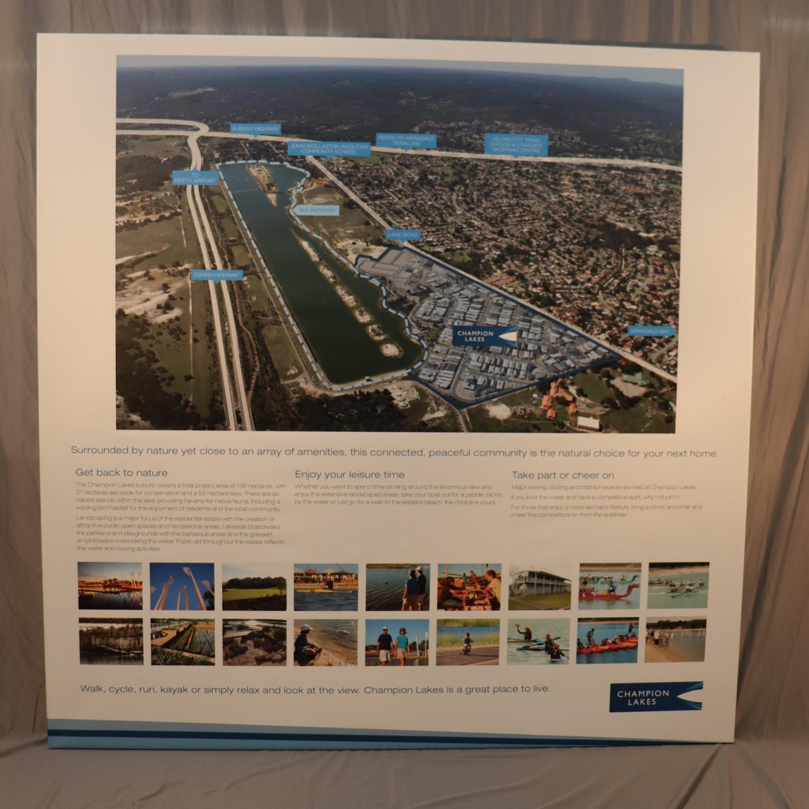 Rectangle white canvas sign, mounted onto a wooden frame. On the top half of the sign is a colour arial photograph of the Champion Lakes Rowing course and to the bottom half features three columns of text with a blue heading over black text with two rows of 18 colour images. 