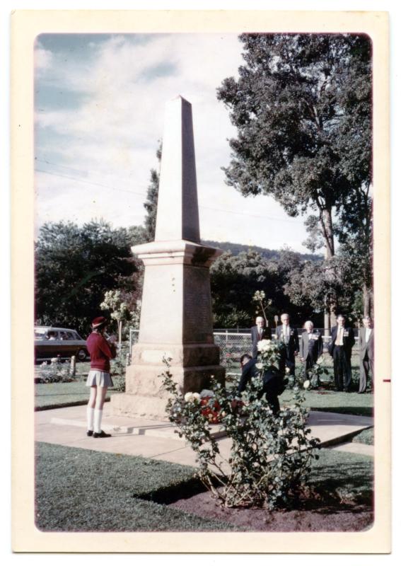 Colour photograph of girl in school uniform standing in front of a stone obelisk shaped war memorial. Five men in background wearing suits and medals