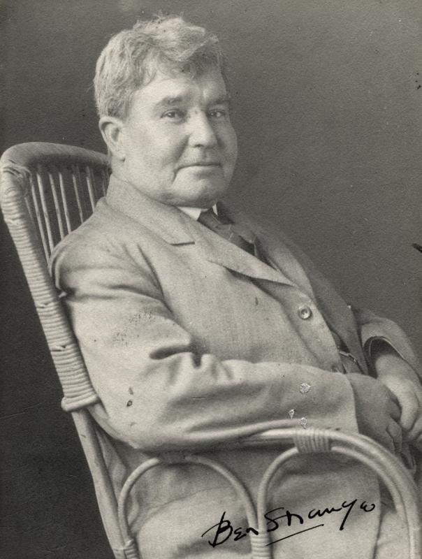 Black and white portrait photo of a man in a jacket sitting in a ratan cane chair
