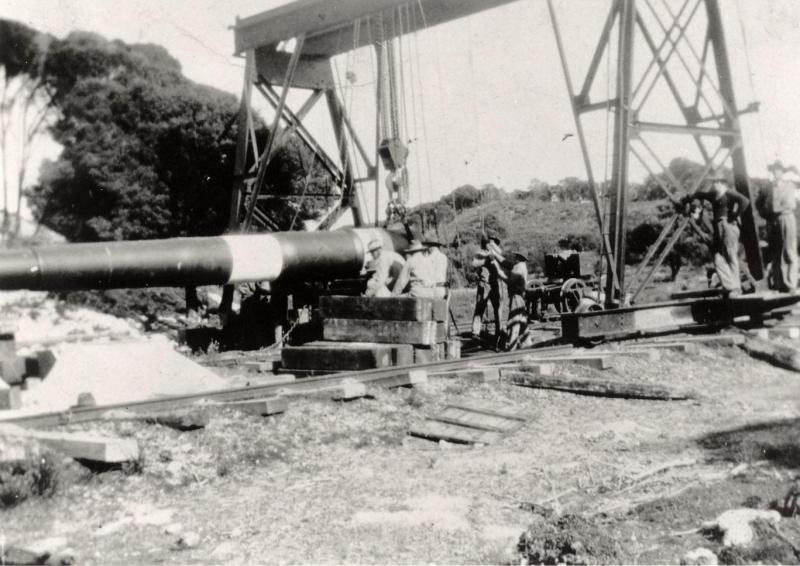 Black-and-white photo showing a group of workers assembling a large 9.2 inch coast artillery weapon outdoors. The barrel is positioned on a platform with scaffolding and machinery around it. The surrounding area includes vegetation and uneven terrain