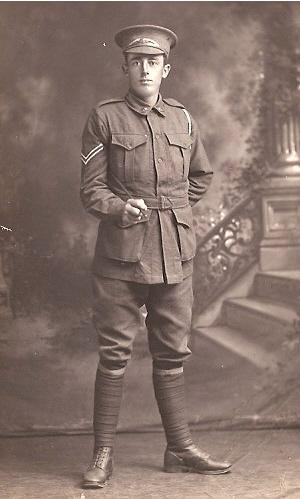 Vintage black-and-white photograph of a soldier in a World War I-era military uniform, standing in a formal pose. The soldier is wearing a peaked cap, jacket, knee-length breeches, and long socks, with a backdrop of decorative stairs and foliage