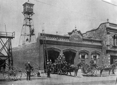 A rectangular brick building labelled 'Fire Station 1904', with two large arched entryways and two arched doorways. A ladder leans against each end of the building. In the foreground the fire brigade pose with equipment. In the background rises a simple triangular observation tower.