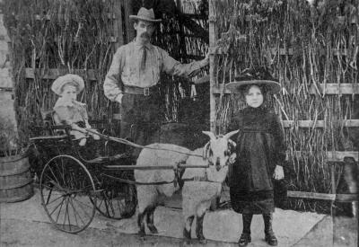 A young girl sits on a goat cart, holding the reigns. An holder girl holds the goat's harness and a man stands behind them, leaning on the doorway of a wooden building.