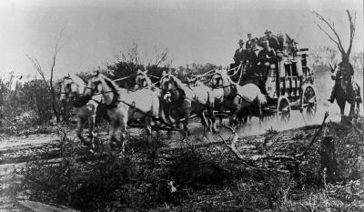 Six harnessed horses pull a coach seating five men along a dusty bush road. A single man armed with a gun rides behind them.