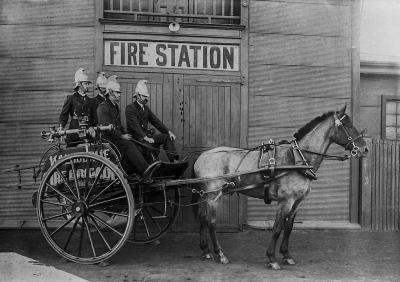 Four fire fighters sit on a small horse-drawn wagon in front of a corrugated iron building labelled 'Fire Station'. The wagon is labelled 'Kalgoorlie Fire Brigade'.