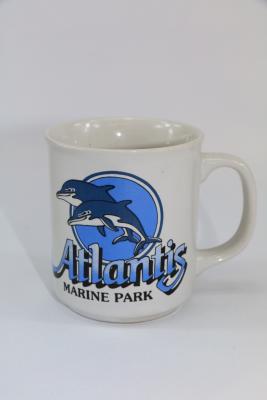  The mug has a printed design in blue and black printed on one side. The design features two jumping dolphins in front of a blue circle with a blue and and white outline. Underneath is the text 'Atlantis MARINE PARK' . Atlantis is written in graduated blue with black shadows and marine park is written in black block letters.