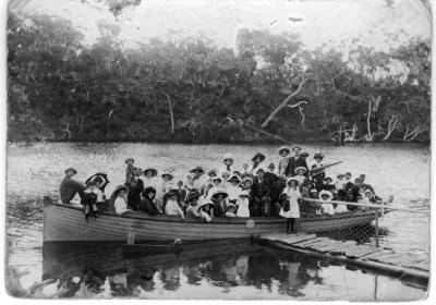 A black and white photographic image of a boat with a large picnic party aboard, the photograph is posed as all the family groups are facing the camera. The fully loaded boat is close alongside on a high tide at a landing jetty on a river