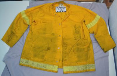 A Yellow firefighter's fire resistant jacket, with yellow fluorescent stripes at the base of the garment, as well as at the bicep area of the sleeves. The jacket is stained with ash.