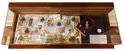 A display case containing various military medals and decorations arranged on a map background. The medals are placed at different points on the map, possibly indicating locations of significance. To the right, there is a blue plaque or document and an additional medal, along with an information card. The display case has a wooden frame with a glass cover.