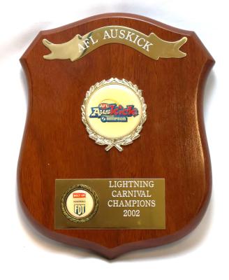 Brown treated and glazed wooden shield shaped plaque with five points. Obverse side has three key features featuring engravings and inscriptions. Along the top is an engraved metal ribbon banner. In the middle there is a raised circular emblem encased in a gilded wreath featuring sponsorship details and AFL Auskick logo. Towards the bottom is a metal rectangle with engraving on the right side and a smaller raised wreath on the left which has extra sponsorship details. 