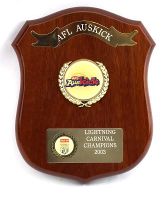 Brown treated and glazed wooden shield shaped plaque with five points. Obverse side has three key features featuring engravings and inscriptions. Along the top is an engraved metal ribbon banner. In the middle there is a raised circular emblem encased in a gilded wreath featuring the AFL Auskick logo. Towards the bottom is a metal rectangle with engraving on the right side and a smaller raised wreath on the left which has extra sponsorship details. 