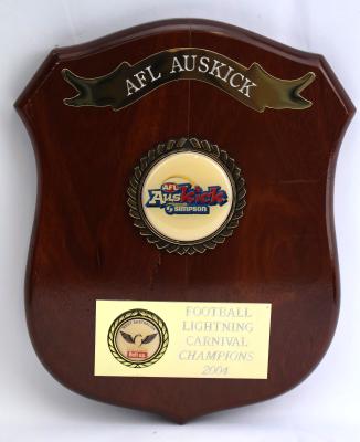 Brown treated and glazed wooden shield shaped plaque with five points. Obverse side has three key features consisting of engravings and inscriptions. Along the top is an engraved metal ribbon banner. In the middle there is a raised circular emblem encased in a golden circular wreath featuring sponsorship details and AFL Auskick logo. Towards the bottom is a metal rectangle with engraving on the right side and a smaller raised wreath on the on the left which has extra sponsorship details. 