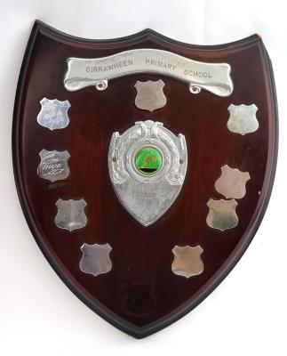 Dark brown treated wooden shield with four points. The obverse side of the shield consists of nine smaller silver shields with each having six points, with only two of the shields featuring engraving of the year and winning faction team. At the top of the shield there is a raised metal ribbon banner that is held in place by two screws and displays the school’s name. Beneath that is one of the smaller metal shields with no engraving. 