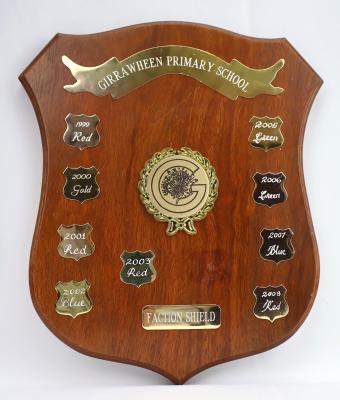 Brown treated wooden shield with four points. The obverse side of the shield consists of nine smaller silver shields with each having six points, with only two of the shields featuring engraving of the year and winning faction team. At the top of the shield there is a raised metal ribbon banner that is held in place by two screws and displays the school’s name. 