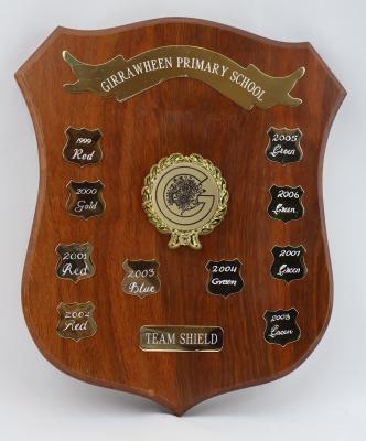 Brown treated wood shield with five points. The obverse side of the shield consists of 10 smaller engraved metal shields featuring team colours and year. At the top of the shield there is a metal ribbon banner. In the middle there is a raised metal wreath with a bow which cocoons around a large letter G and floral emblem. Towards the bottom of the shield is another smaller rounded rectangle that is also engraved.