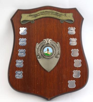 Brown treated wooden shield shaped plaque with five points. The obverse side consists of 11 small silver shields, some of which have been engraved with the year and winning faction colour. There are five shields on the left side and six on the right side. At the top of the shield is a raised metal ribbon banner detailing the name of the school and sporting event. In the middle of the shield is a raised detailed metal shield with three points.