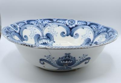 A large, ceramic bowl with a scalloped rim. The inside of the bowl is white, painted with ornate blue detailing. The outside of the bowl is similarly painted with flowers and leaves in three places. Cracks from outside edge down to bottom. There is some black marks on one of the edges.
