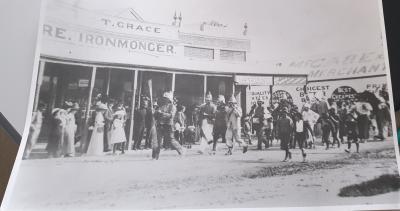 Large black and white photograph Possibly 1900's - 1910's.  Kellerberrin Fire Brigade leading parade past McCabes Merchants Store, T Grace - Iron Monger store Massingham St