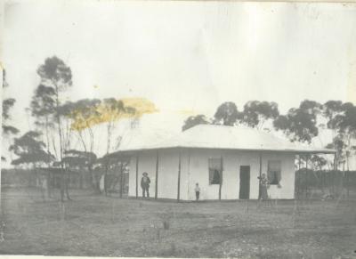 Black and White photograph.  Laird homestead with 3 people, 2 adults and 1 child, on verandah.